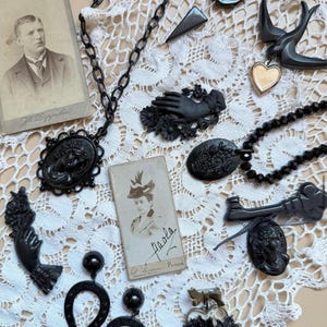 May include: A collection of black jewellery including a cameo necklace, hand-shaped brooches, horseshoe earrings, and a heart pendant, displayed alongside two vintage sepia-toned portrait photographs on a white lace fabric background.