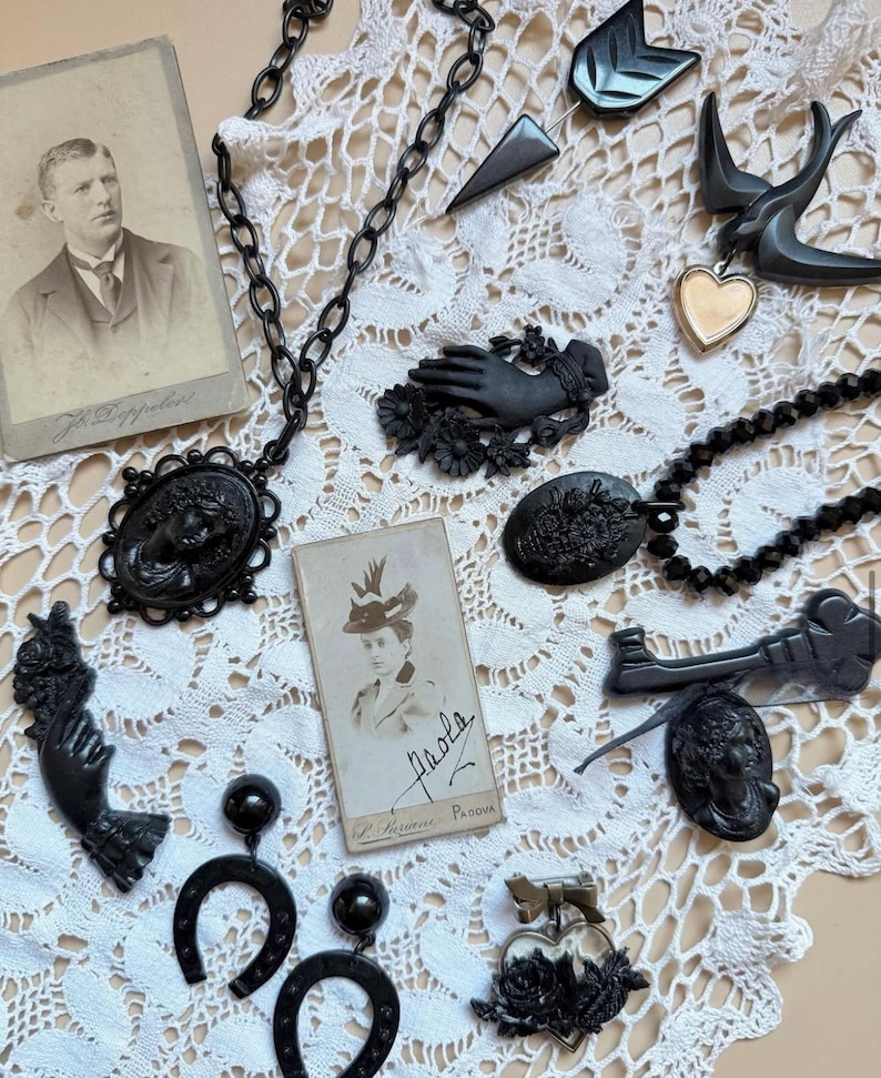 May include: A collection of black jewellery including a cameo necklace, hand-shaped brooches, horseshoe earrings, and a heart pendant, displayed alongside two vintage sepia-toned portrait photographs on a white lace fabric background.