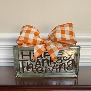 May include: A glass block with the words "Happy Thanksgiving" in brown vinyl lettering. The block is decorated with a large orange and white gingham bow.