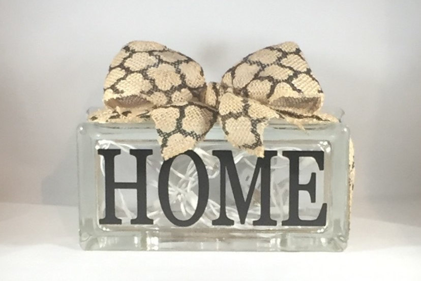 May include: A clear glass block with the word "HOME" in black lettering. A decorative bow made of tan burlap with a brown geometric pattern sits atop the block. White fairy lights are visible inside the block.