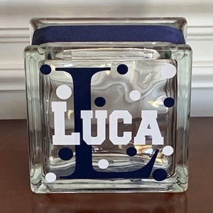 Pu&ograve; includere: Un blocco di vetro trasparente con un nastro blu navy attorno alla parte superiore. Il blocco ha un motivo a pois bianchi e una lettera "L" blu navy con il nome "Luca" in lettere bianche.