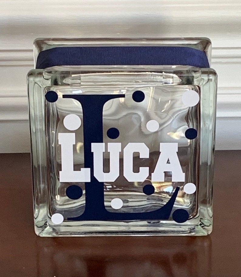 Pu&ograve; includere: Un blocco di vetro trasparente con un nastro blu navy attorno alla parte superiore. Il blocco ha un motivo a pois bianchi e una lettera "L" blu navy con il nome "Luca" in lettere bianche.