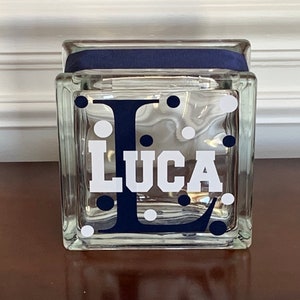 Pu&ograve; includere: Un blocco di vetro trasparente con un nastro blu navy attorno alla parte superiore. Il blocco ha pois bianchi e una lettera "L" blu navy con il nome "Luca" in lettere bianche.