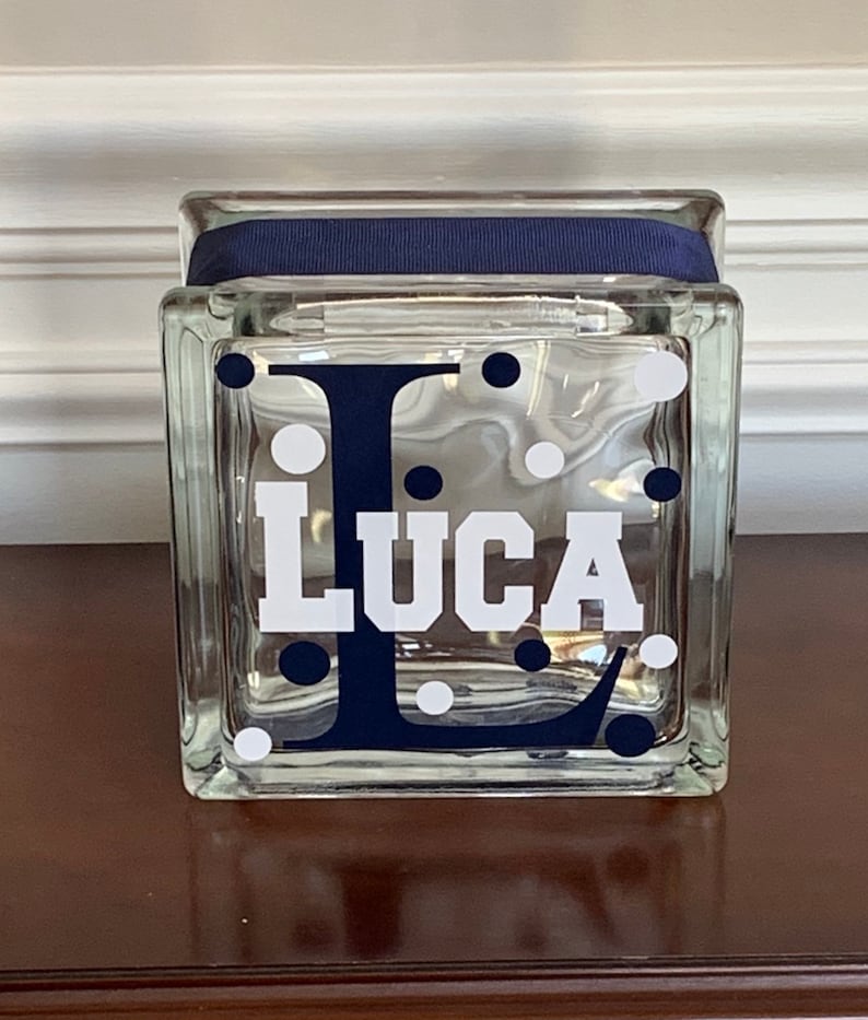 Pu&ograve; includere: Un blocco di vetro trasparente con un nastro blu navy attorno alla parte superiore. Il blocco ha pois bianchi e una lettera "L" blu navy con il nome "Luca" in lettere bianche.