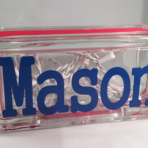 May include: A clear glass block with a red ribbon around the top. The block contains a string of white lights and a blue vinyl decal that says "Mason".