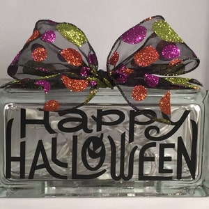 May include: A clear glass block with the words "Happy Halloween" in black script. The block is topped with a black sheer bow adorned with glittery orange, green, and purple polka dots. The block is lit with white lights.