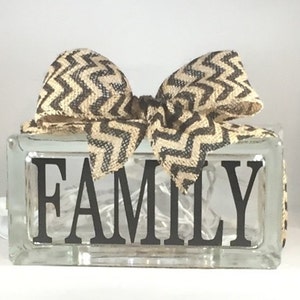 May include: A glass block with the word "FAMILY" in black lettering, decorated with a brown and beige chevron patterned bow. The block is illuminated with white fairy lights.
