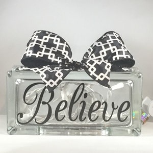 May include: A glass block with a black and white geometric patterned ribbon tied around it. The block has a string of white lights inside and the word "Believe" is written on the front in black.