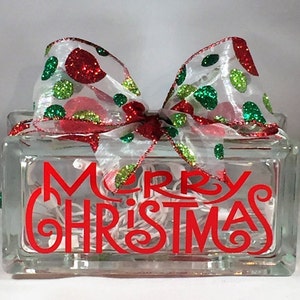 May include: A glass block with the words "Merry Christmas" in red lettering. The block is decorated with a white ribbon with red and green glitter polka dots.