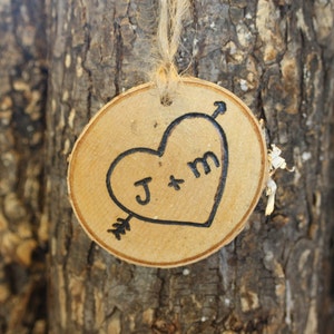 Op de afbeelding: Een houten hartvormige hanger met de initialen "J & M" erin gegraveerd. Het hart wordt doorboord door een pijl en hangt aan een touwtje.