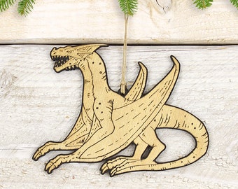 May include: A wooden dragon ornament with a detailed etched design. The dragon is light brown with dark brown etched lines, wings, and a long tail. It hangs from a natural twine string, with evergreen branches in the background.