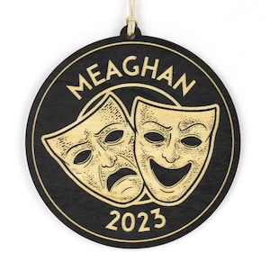 May include: A round, black wooden ornament with the name "MEAGHAN" in a circular font. The ornament features engraved comedy and tragedy masks and the year "2023" in a light wood color. A string is attached for hanging.
