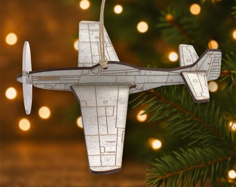 Könnte beinhalten: Ein Holzflugzeug-Ornament mit Propeller und Aufhängeschnur. Das Ornament ist in Grau- und Brauntönen bemalt, mit detaillierten Linien, die die Merkmale des Flugzeugs darstellen. Der Hintergrund ist verschwommen mit Tannenzweigen und Lichtern.