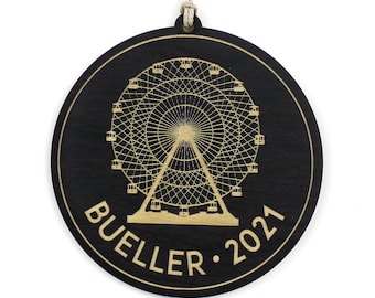 May include: A round, black wooden ornament with a Ferris wheel design in gold. The text "BUELLER 2021" is etched around the edge. A string is attached for hanging. The ornament is suitable for holiday decor.
