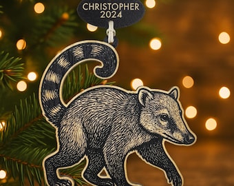 Könnte beinhalten: Weihnachtsbaumschmuck aus Holz mit einer detaillierten Gravur eines Nasenbären, mit gestreiftem Schwanz und langer Schnauze. Das Ornament hat ein kleines, schwarzes Etikett mit dem Namen "CHRISTOPHER" und dem Jahr "2024".