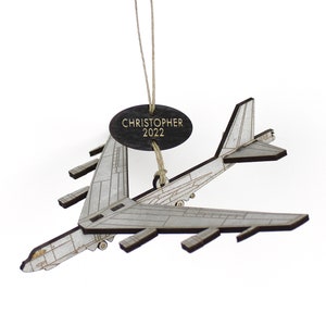 May include: A wooden airplane ornament with a silver and brown finish. The ornament has the name "CHRISTOPHER" and the year "2022" on a dark oval tag. The plane is suspended by a natural twine.