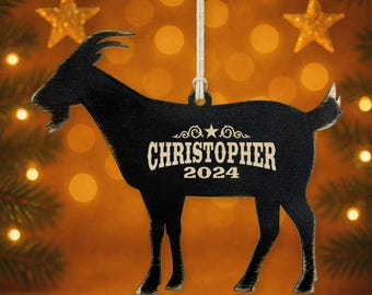 May include: Black goat-shaped Christmas ornament with the name "CHRISTOPHER" and the year "2024" in white lettering. The ornament hangs from a white string, with two gold star ornaments in the background.