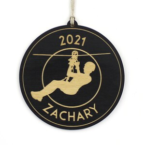 May include: A round, black wooden ornament with a light wood-colored design. The design features a person ziplining, the year "2021", and the name "ZACHARY". The ornament hangs from a natural-colored string.