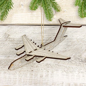May include: A wooden airplane ornament with a light gray body and brown trim, suspended by a natural twine string. The ornament features detailed cutouts of the plane's wings, tail, and fuselage. Green pine branches are in the background.