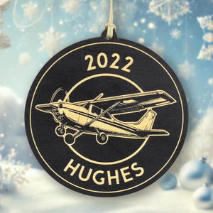 May include: A round, black wooden Christmas ornament with a gold airplane design. The ornament has the year "2022" at the top and the name "HUGHES" at the bottom. A string is attached for hanging.