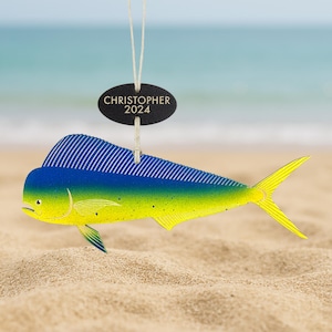 May include: A colorful fish ornament with a blue and yellow design, hanging by a string. The ornament has a fin with blue stripes and a yellow tail. A black oval tag above reads "CHRISTOPHER 2024". The ornament is on a sandy beach.