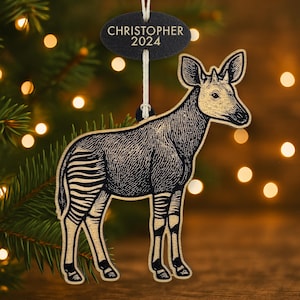 May include: Wooden okapi ornament with black and tan details. The ornament features a detailed illustration of an okapi with striped legs and the text "CHRISTOPHER 2024" on a black tag. The ornament hangs from a white string.