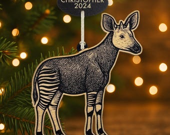 Könnte beinhalten: Holz-Okapi-Ornament mit schwarzen und beigen Details. Das Ornament zeigt eine detaillierte Illustration eines Okapi mit gestreiften Beinen und dem Text "CHRISTOPHER 2024" auf einem schwarzen Anhänger. Es hängt an einer weißen Schnur.
