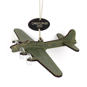 May include: A wooden olive green airplane ornament with clear propellers. The ornament has the name "CHRISTOPHER" and the year "2022" engraved on a dark brown oval tag. The airplane is a detailed model.