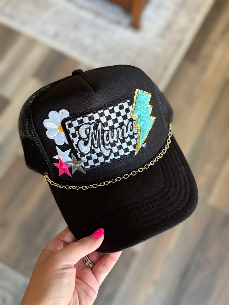 May include: Black trucker hat with a checkered patch that says "Mama" in white script. The hat also has a blue lightning bolt patch, a white daisy patch, and two pink and silver star patches. A gold chain is attached to the hat.