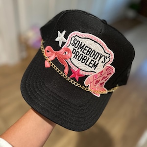 May include: Black trucker hat with a pink and white patch that says "Somebody's Problem" and a pink bow, star, and boot embellishments.