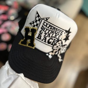 May include: Black and white trucker hat with a white crown and black bill. The hat has a black and white checkered star, a black and white butterfly, and a white patch with the text "Expensive Difficult & Talk Back" on it.