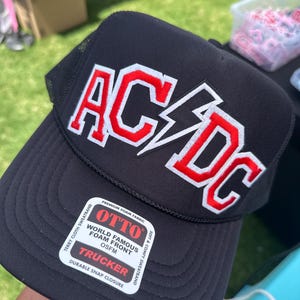 May include: Black trucker hat with a red and white AC/DC logo. The hat has a mesh back and a snap closure. The hat is made by Otto and features a foam front.