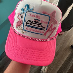 May include: A hot pink trucker hat with a white front panel. The hat features embroidered patches including a flamingo, a starfish, and the text "I Like Big Boats & I Can Not Lie" with a boat graphic. A silver chain accents the brim.