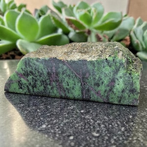 Pode incluir: Uma pedra bruta com um padrão salpicado verde e preto, acentuado por raias roxas. A borda superior tem uma superfície áspera, texturizada e castanha clara. A pedra é exibida em uma superfície escura e salpicada.
