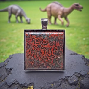 May include: Square pendant with a red and black patterned stone set in a silver-tone metal frame. The pendant is displayed on a dark, textured surface, with toy dinosaurs in the background on green grass.