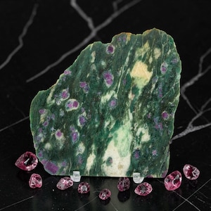May include: A flat, irregularly shaped slab of green zoisite mineral featuring embedded magenta ruby crystals, displayed upright on a small clear stand, surrounded by several loose, faceted magenta ruby gemstones on a dark surface.
