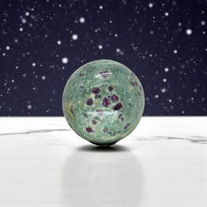 May include: A green sphere with red and purple inclusions, resting on a white surface. The gemstone ball has a speckled appearance and is set against a dark blue background with white dots.