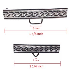 Pode incluir: Duas fivelas de cinto de metal prateado com um design de elos de corrente. A fivela superior tem 4,1 cm de comprimento e a fivela inferior tem 3,2 cm de comprimento. Cada fivela tem um pequeno laço para fixação.