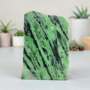 May include: A rectangular, rough-edged stone with a vibrant green hue, streaked with bold black veins. The natural stone is standing upright, showcasing its unique pattern and texture. The stone's surface has a slightly uneven texture, adding to its organic appeal.