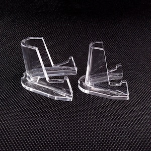 May include: Two clear plastic display stands for collectibles. The stands are shaped like a small, angled platform with a raised back.