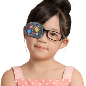 May include: A gray eye patch with colorful embroidered flowers, a sun, and other designs. The eye patch is worn by a young person with dark hair and glasses.