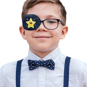 May include: A child wearing a white shirt with a navy blue bow tie and suspenders. The child is wearing a black eye patch with a yellow star and two eyes embroidered on it.