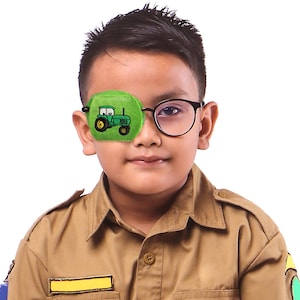 May include: A green eye patch with a cartoon tractor design. The eye patch is being worn by a child wearing glasses.