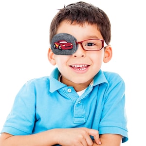 May include: A young boy wearing red-framed glasses and a grey eye patch with a red race car design. The eye patch is made of soft felt.