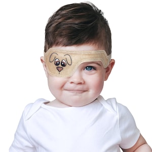 May include: A beige eye patch with a cartoon dog face embroidered on it, worn by a baby. The eye patch is secured with a strap that goes around the head. The baby is wearing a white onesie.