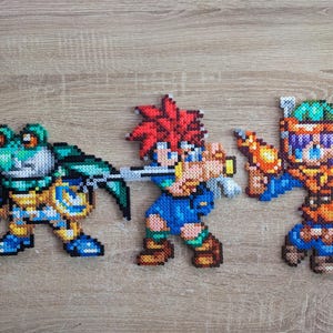 Peut inclure: Trois figurines en pixel art faites de perles en plastique colorées, représentant des personnages d'un jeu vidéo. Elles comprennent un guerrier grenouille avec une épée, un personnage roux avec une épée et un personnage aux cheveux violets avec une arme.