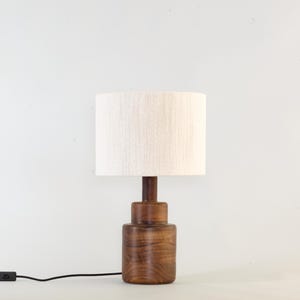Puede incluir: Una l&aacute;mpara de mesa de madera con base cil&iacute;ndrica y pantalla color crema. La l&aacute;mpara tiene un acabado marr&oacute;n oscuro y un cable de alimentaci&oacute;n negro con un interruptor de encendido/apagado. La l&aacute;mpara mide unos 30 cm de alto.
