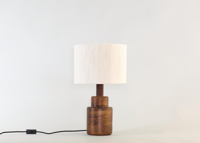 Puede incluir: Una l&aacute;mpara de mesa de madera con base cil&iacute;ndrica y pantalla color crema. La l&aacute;mpara tiene un acabado marr&oacute;n oscuro y un cable de alimentaci&oacute;n negro con un interruptor de encendido/apagado. La l&aacute;mpara mide unos 30 cm de alto.