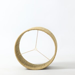 Puede incluir: Una pantalla de l&aacute;mpara beige envuelta en cuerda de color natural. El marco circular tiene tres varillas de soporte doradas que se extienden desde un punto central. La pantalla est&aacute; dise&ntilde;ada para colocarse en una base de l&aacute;mpara.