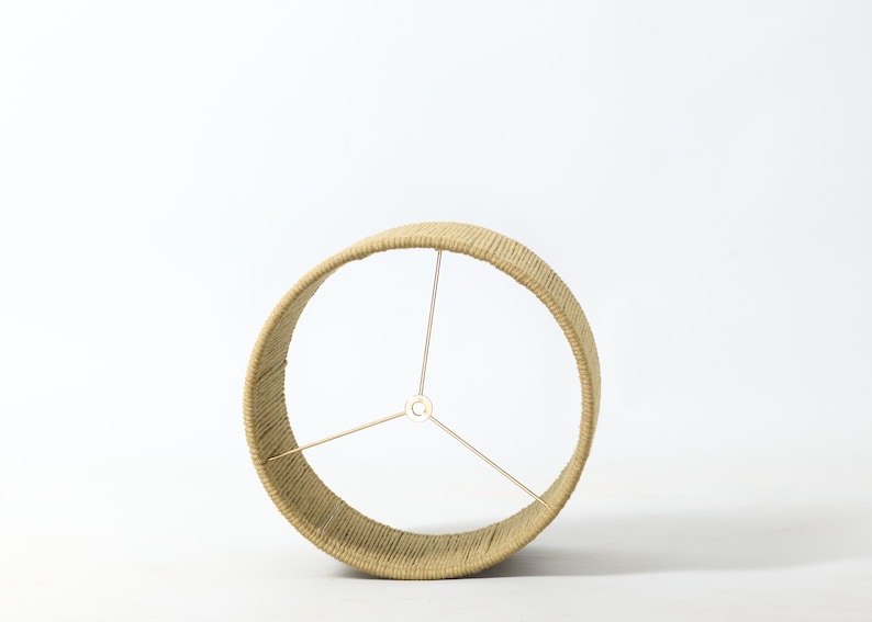 Puede incluir: Una pantalla de l&aacute;mpara beige envuelta en cuerda de color natural. El marco circular tiene tres varillas de soporte doradas que se extienden desde un punto central. La pantalla est&aacute; dise&ntilde;ada para colocarse en una base de l&aacute;mpara.